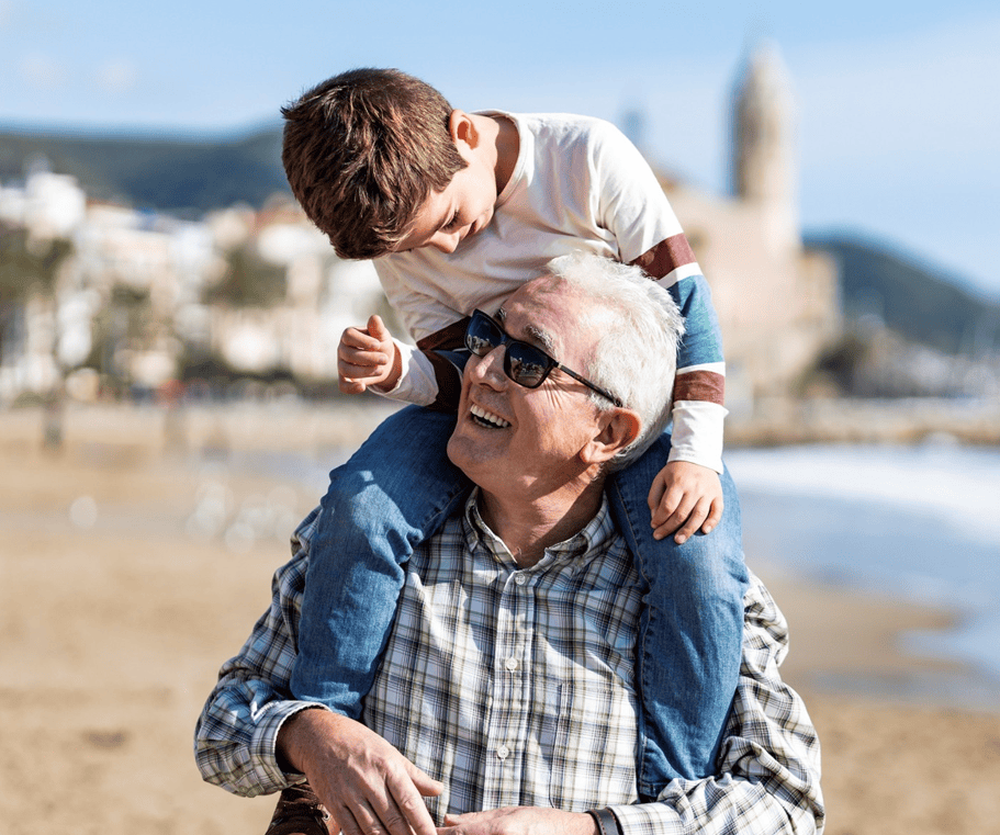 Grandfather carrying grandson on his shoulders on a beach, both smiling, coastal town in the background.