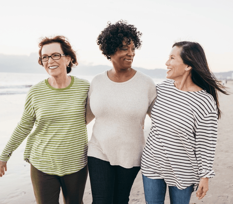 Three women friends smiling and walking arm in arm on the beach.