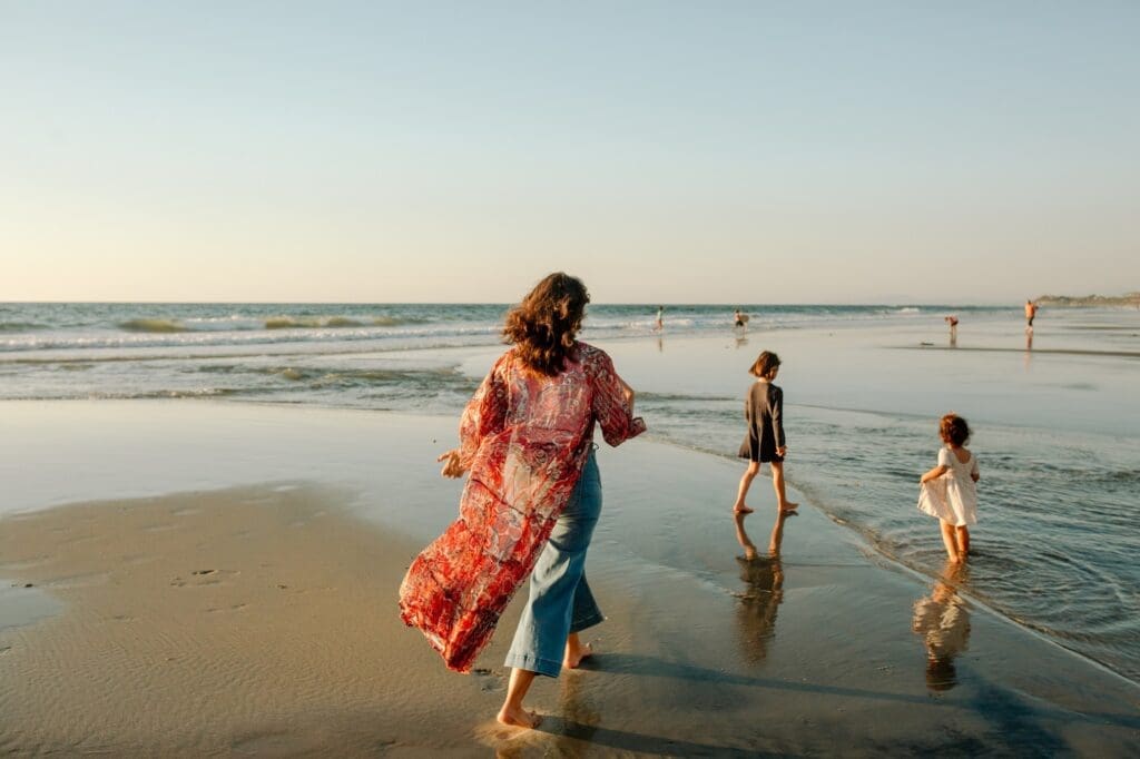 Woman in a patterned kimono walking along the shoreline with two children at sunset.